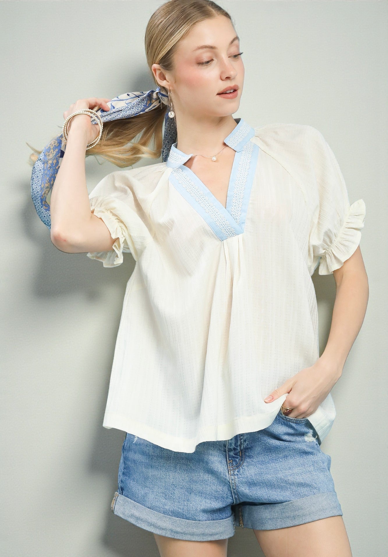 Woman wearing a white blouse with blue trim and denim shorts against a gray background
