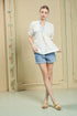 Woman wearing a white blouse with a blue collar and denim shorts in a minimal indoor setting.