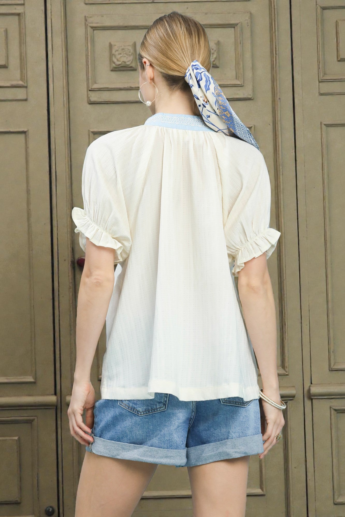 Woman wearing a white blouse and blue shorts with a headscarf, standing in front of a wooden door.