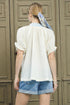 Woman wearing a white blouse and blue shorts with a headscarf, standing in front of a wooden door.