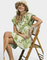 Woman in a green and white dress with a straw hat sitting on a wooden chair against a white background