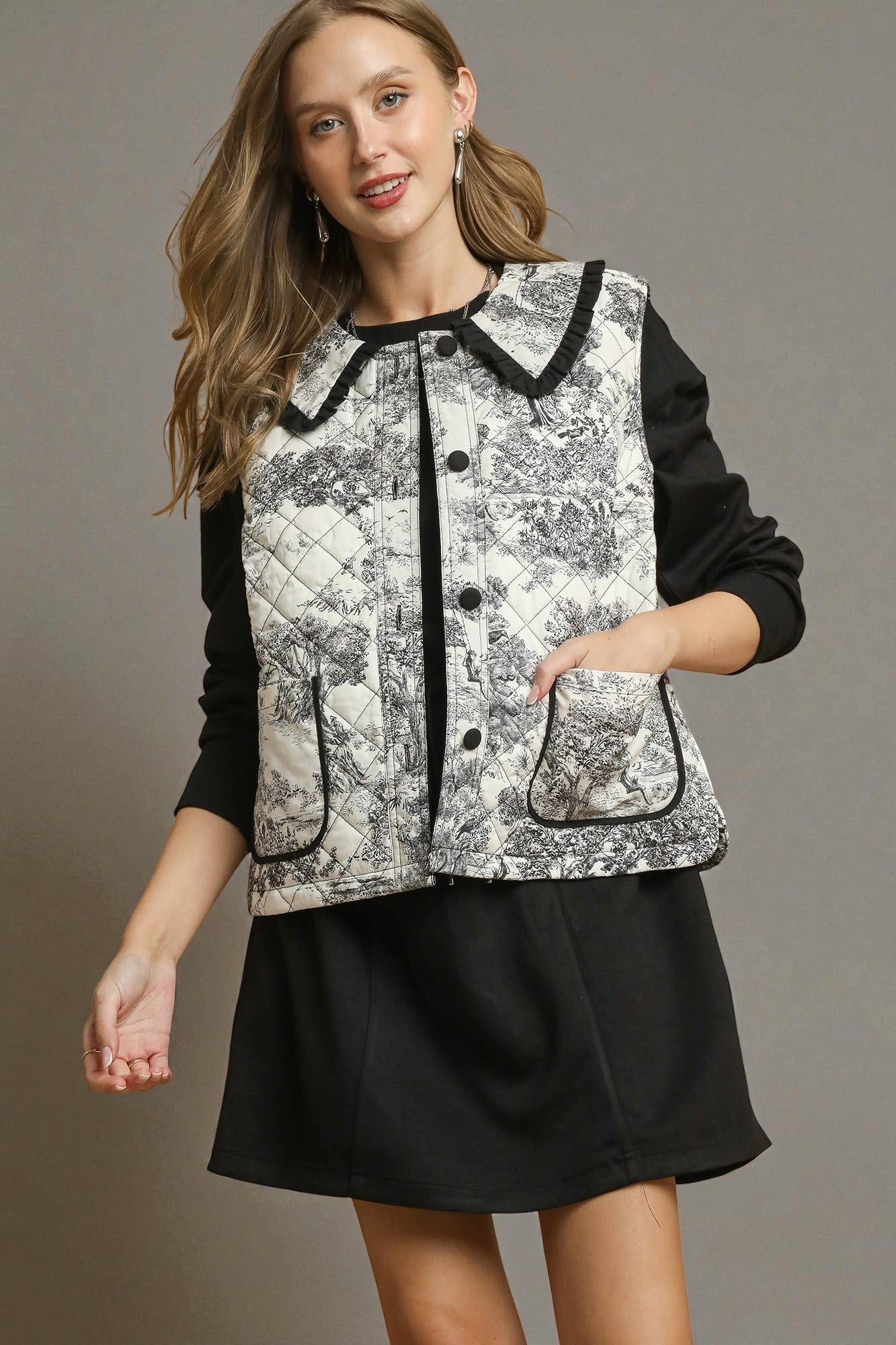 Woman wearing a black and white patterned vest over a black long-sleeve shirt and skirt on a gray background