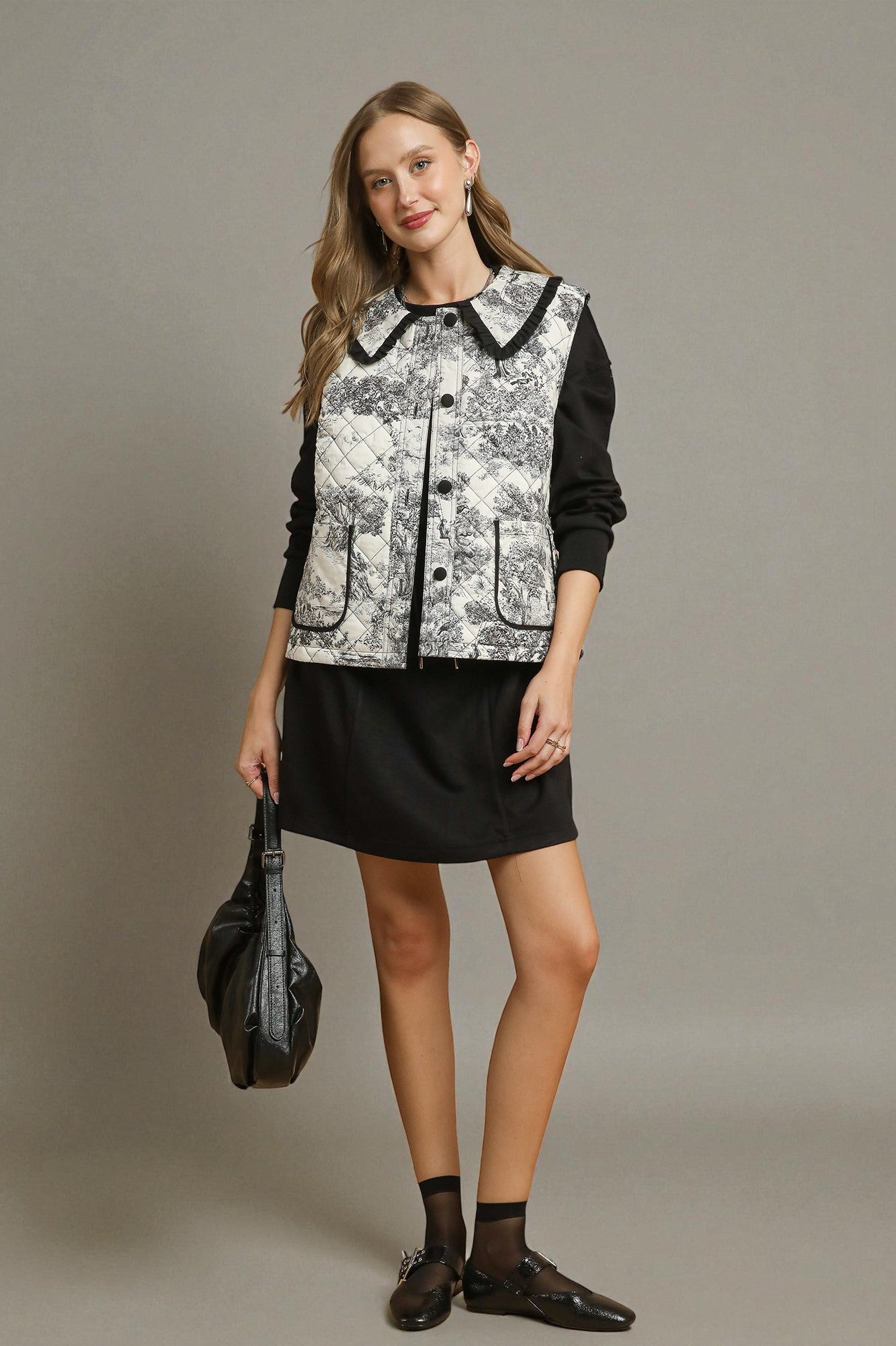 Woman wearing a patterned vest over a black shirt with a black skirt, holding a black bag on a gray background.