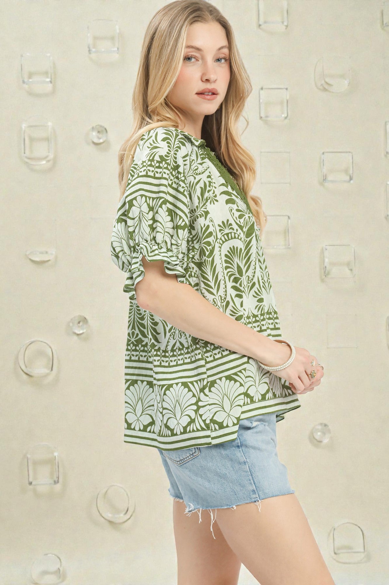 Woman wearing a green patterned top and denim shorts against a neutral background