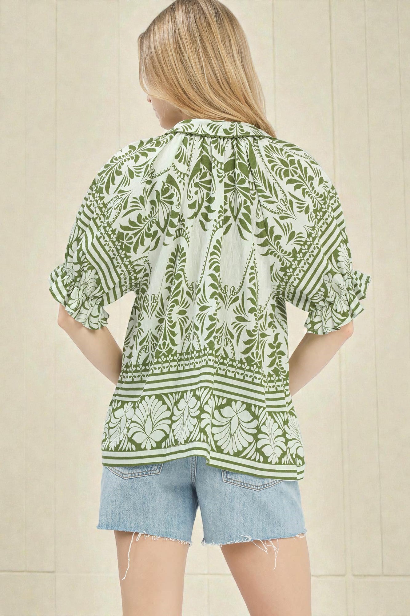 Woman wearing a green and white patterned blouse with denim shorts against a beige wall.