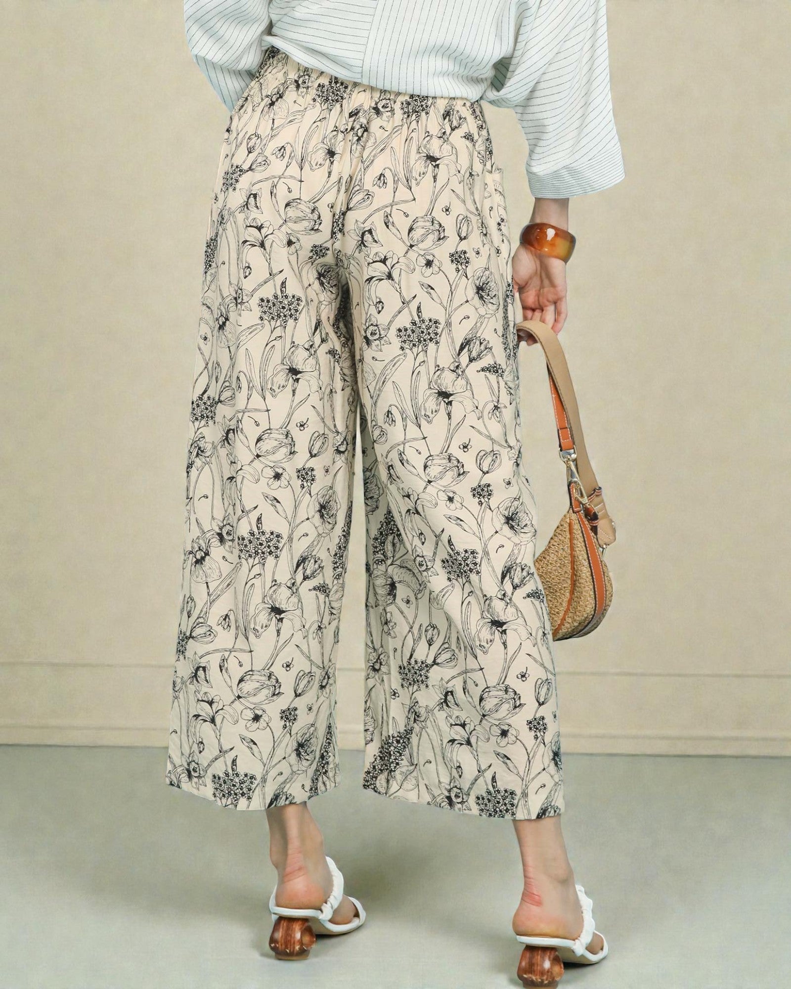 Person wearing floral pants and a striped shirt, holding a woven bag on a neutral background