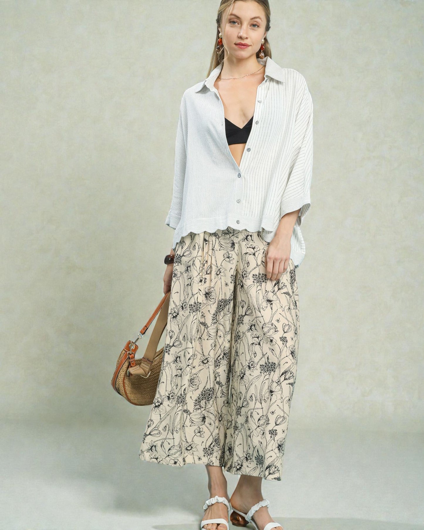 Woman wearing a white shirt, black top, and patterned pants with a beige bag on a light background