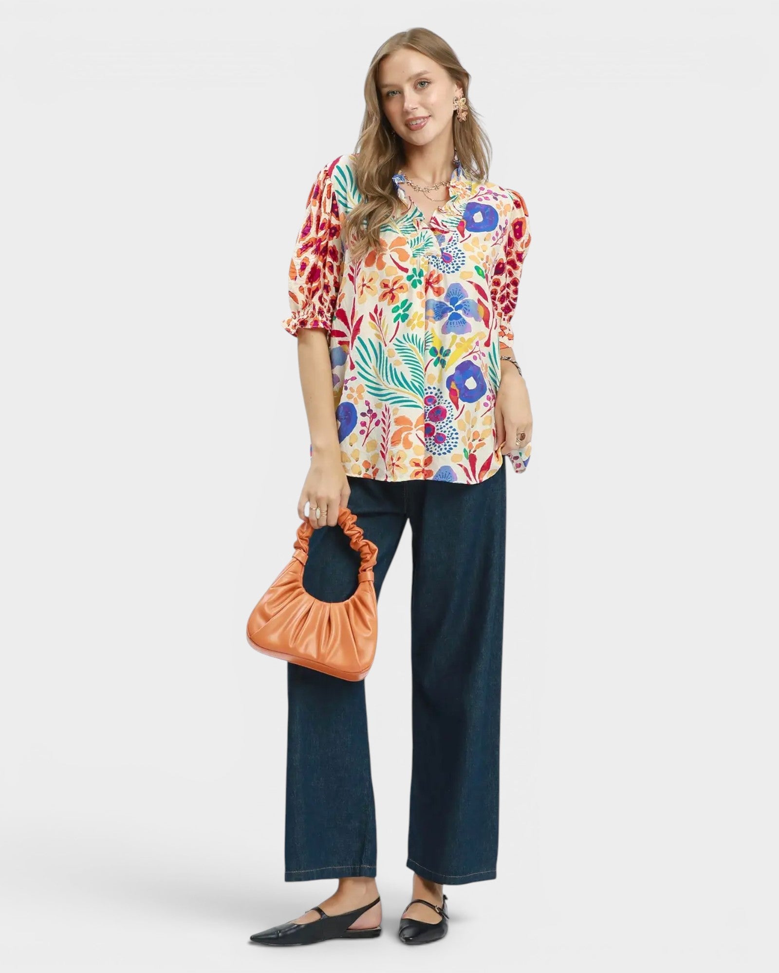 Woman wearing a colorful floral blouse and dark pants, holding an orange bag on a white background