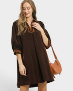 Woman wearing a brown dress with embroidery, holding a brown handbag on a white background