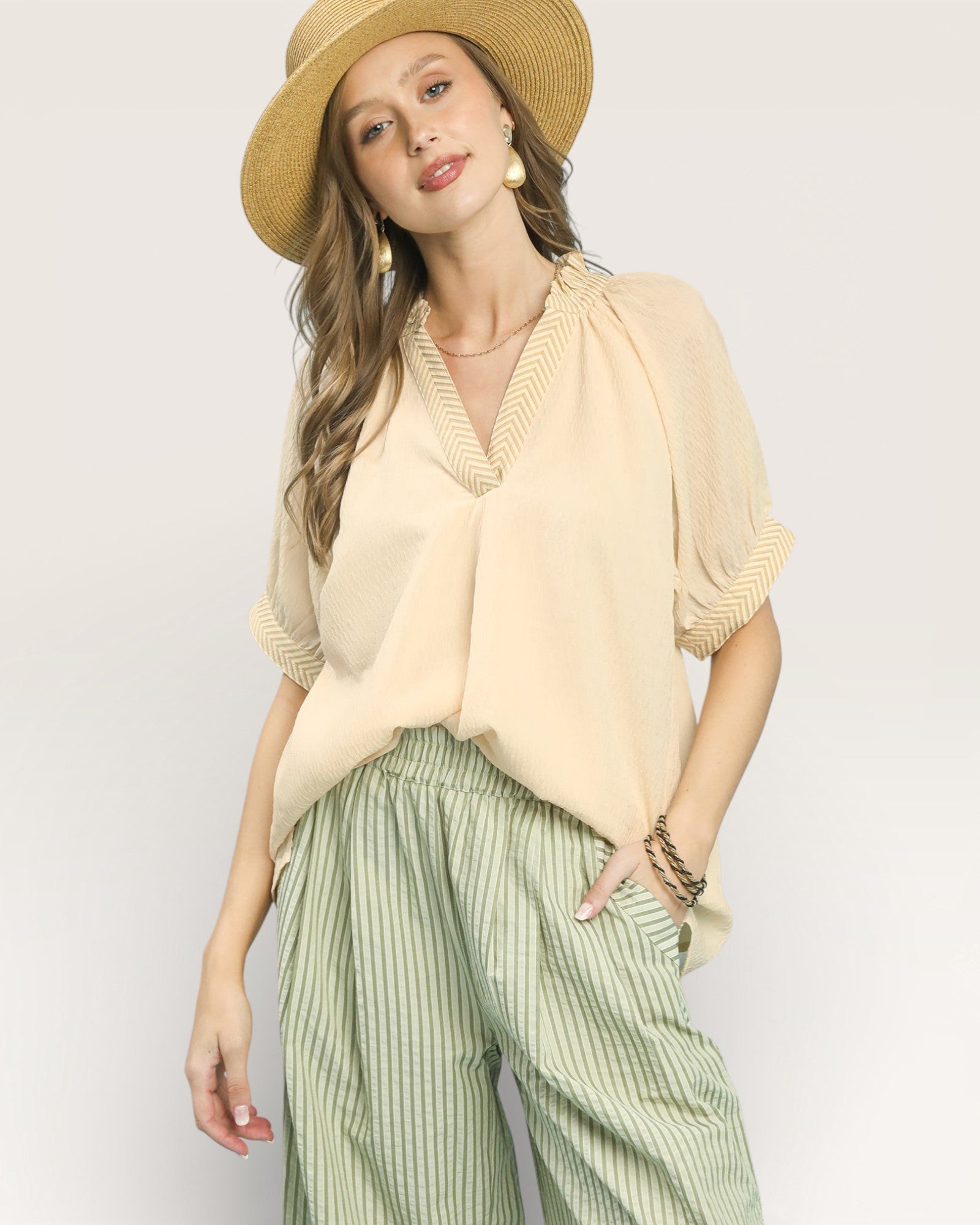 Woman wearing a beige top, green striped pants, and a straw hat on a light background