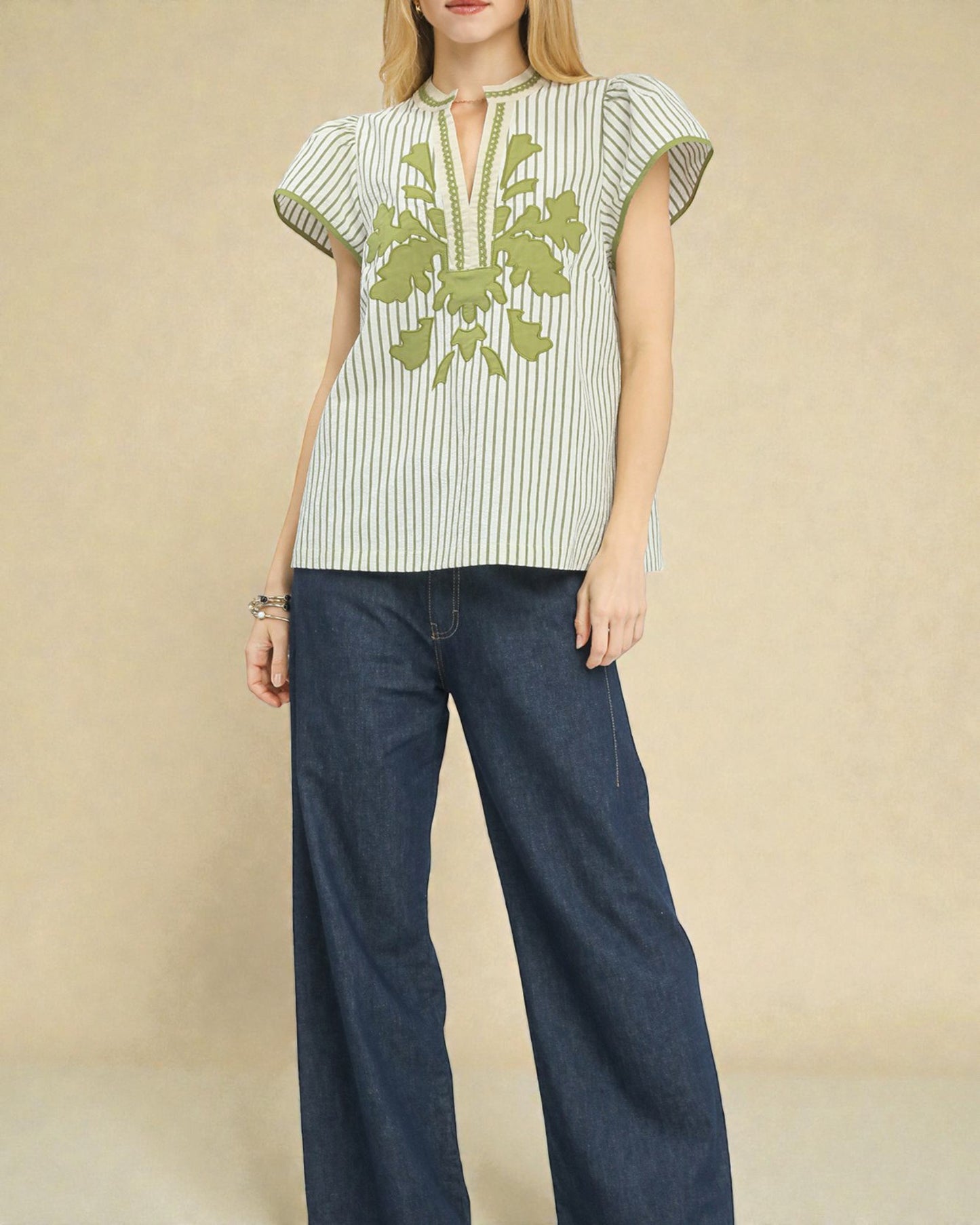Woman wearing a green and white striped top with floral embroidery and blue jeans on a beige background