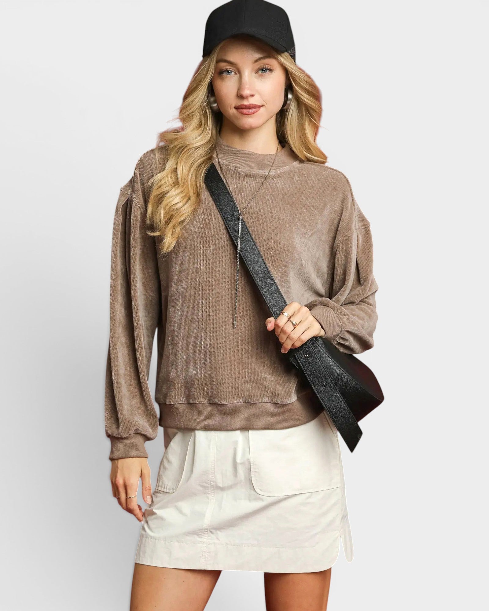 Woman wearing a brown sweatshirt and white skirt holding a black bag against a plain background