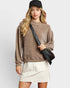 Woman wearing a brown sweatshirt and white skirt holding a black bag against a plain background