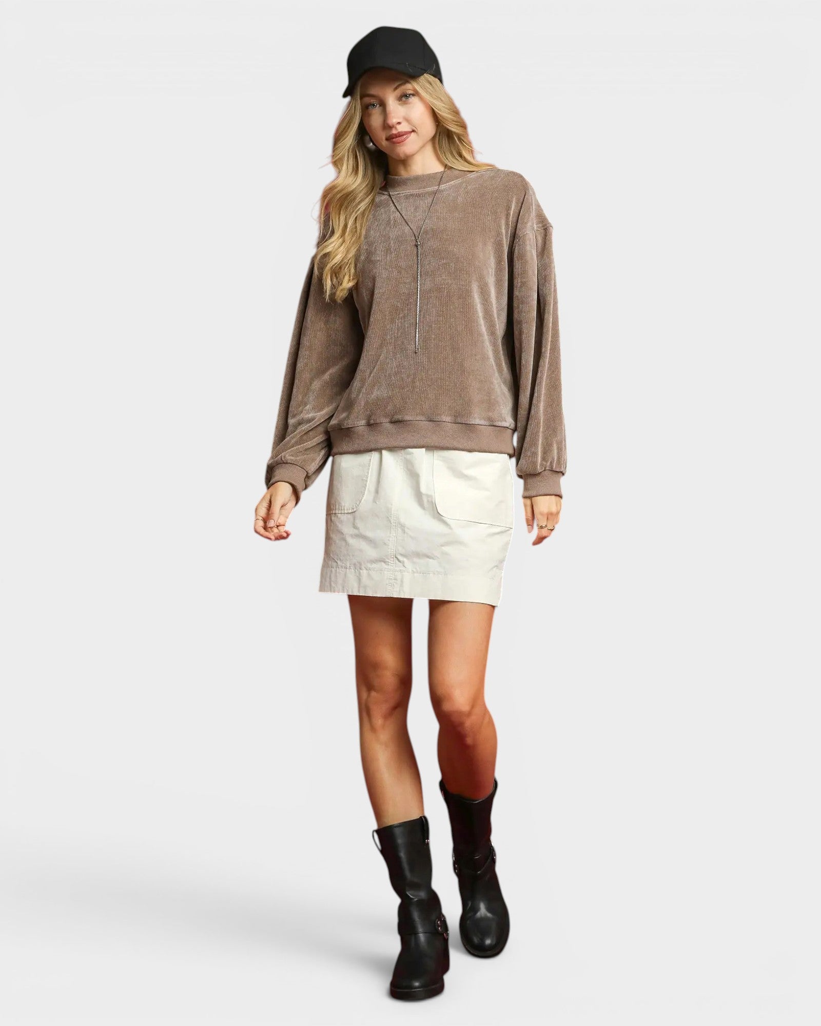 Woman wearing a brown sweatshirt, white skirt, and black boots on a plain background