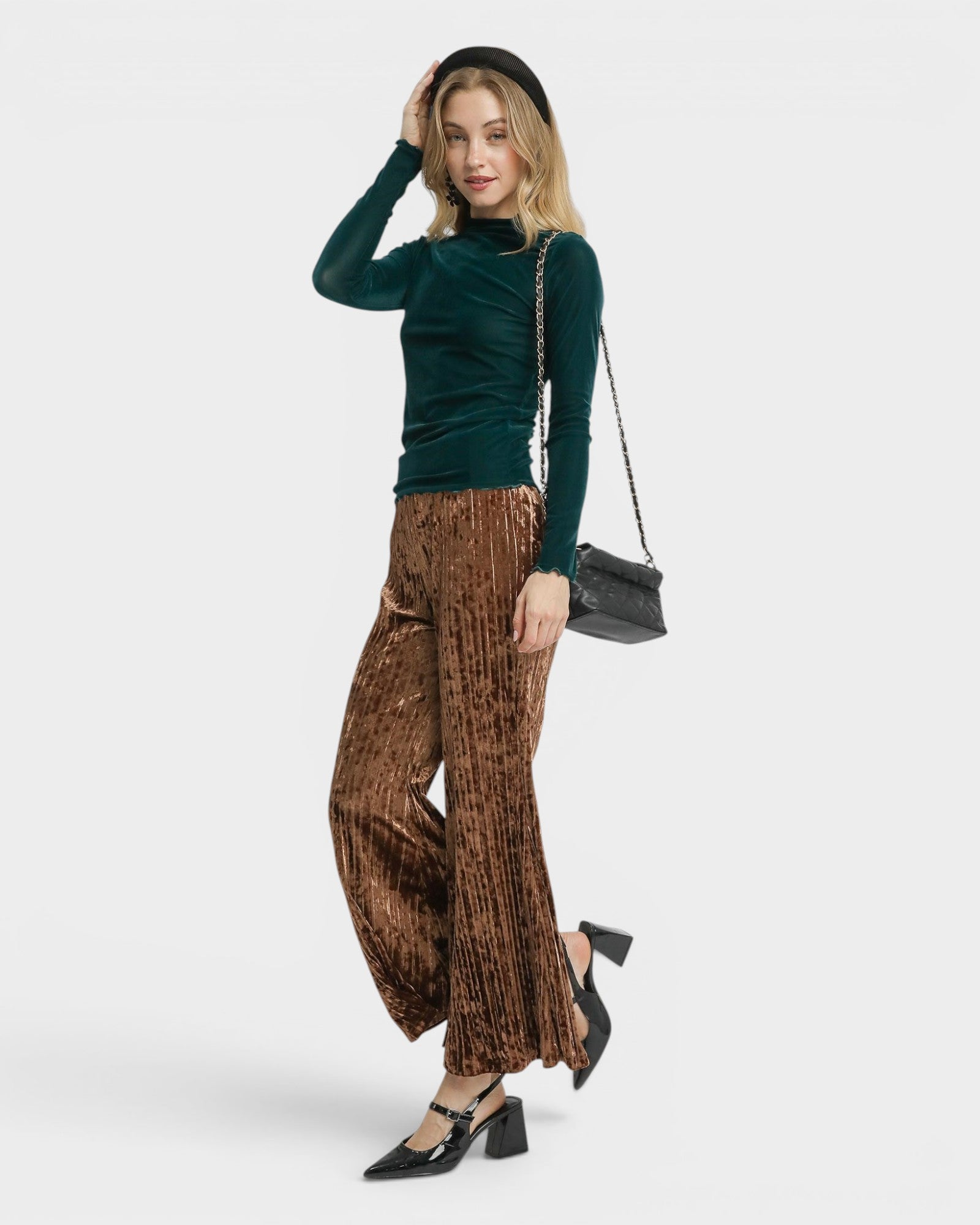 Woman wearing a green velvet top and brown patterned pants with a black handbag on a white background