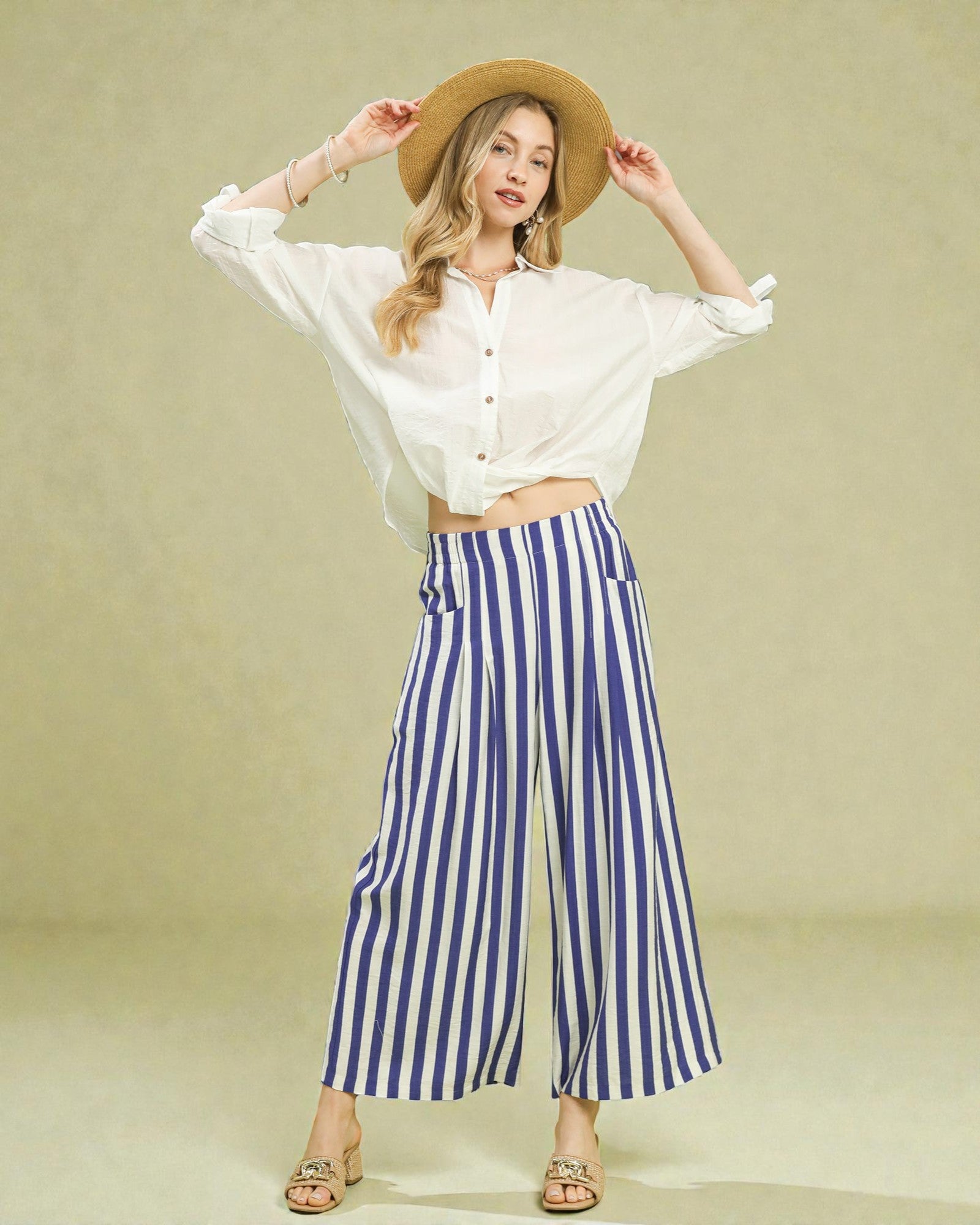 Woman wearing a white shirt, blue striped pants, and a straw hat on a beige background