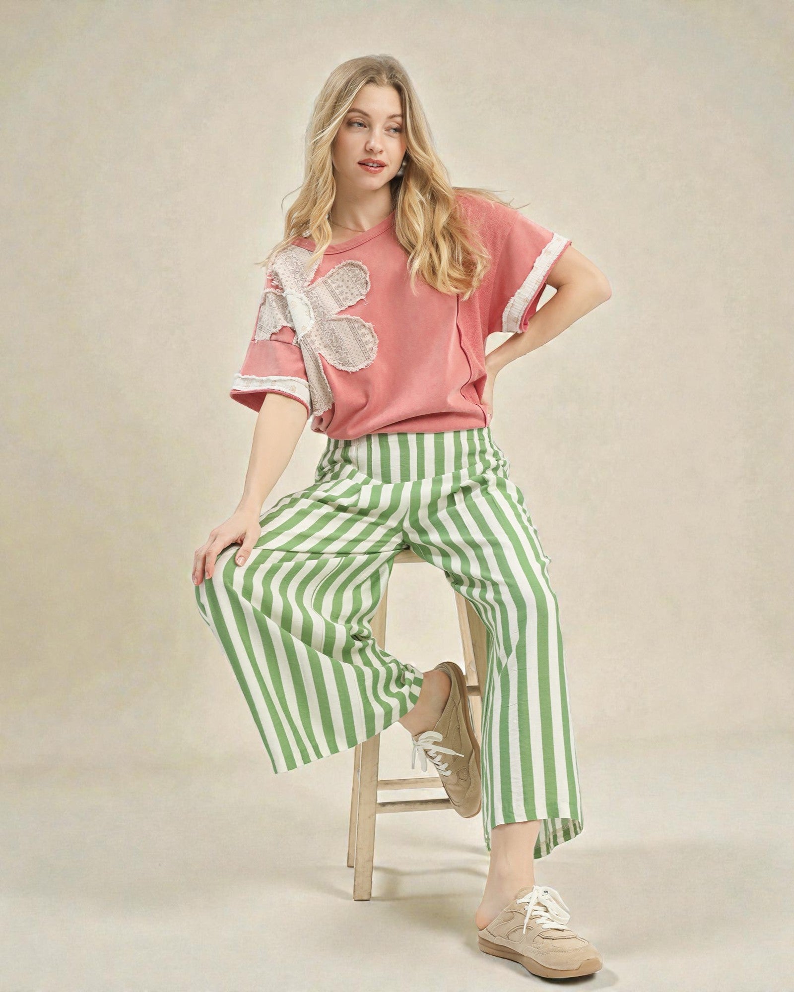Woman wearing a pink top with a white butterfly design and green striped pants, sitting on a wooden stool.