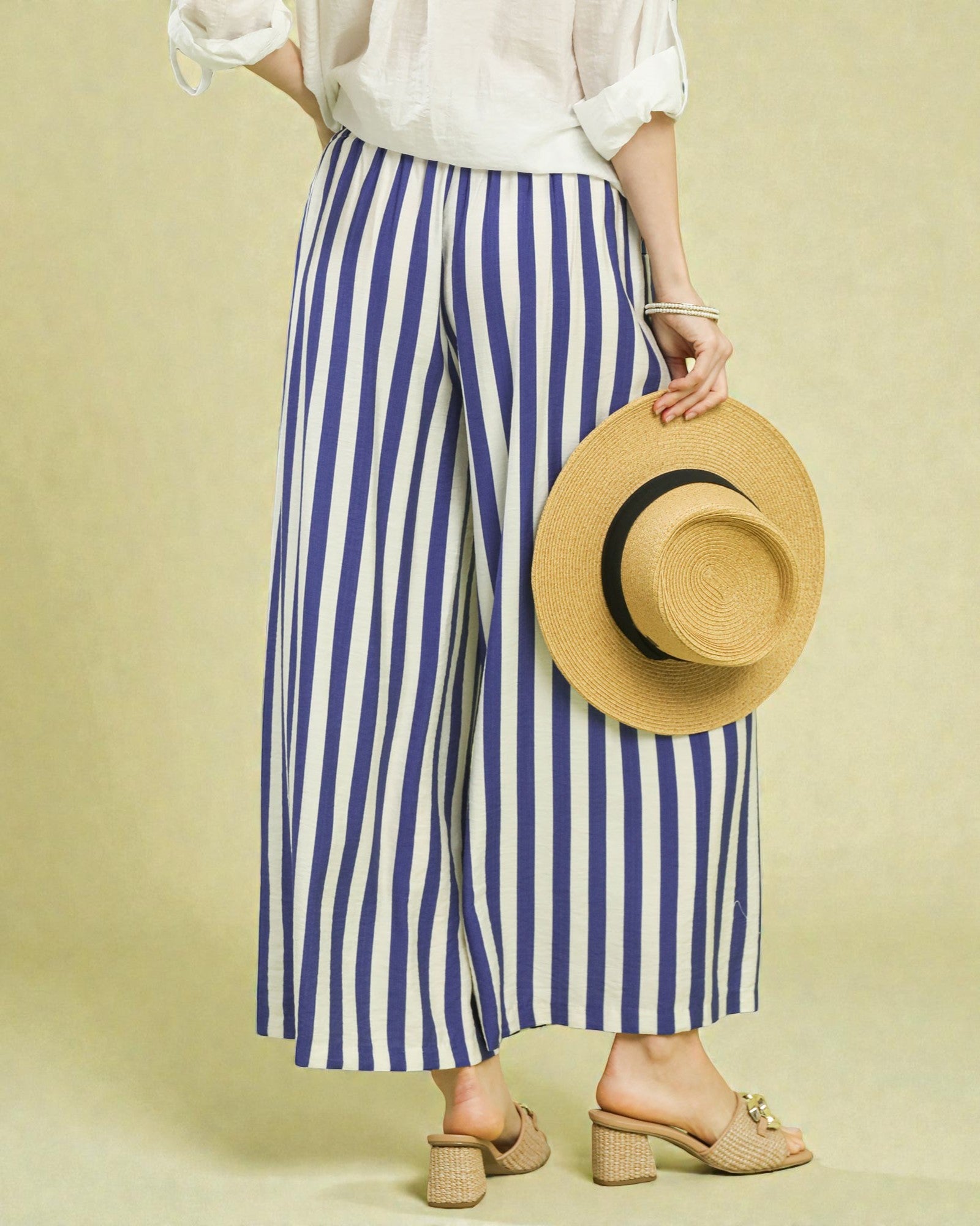 Person wearing blue and white striped pants holding a straw hat on a beige background
