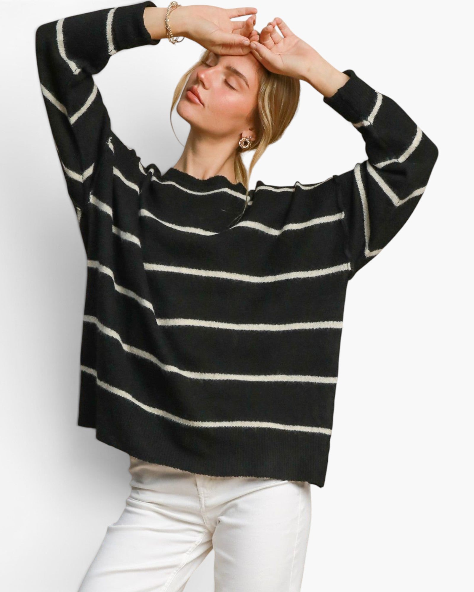 A woman wearing a black knit sweater with horizontal cream stripes and white pants, posing with her hands on her head against a plain white background.