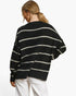 A woman shown from the back wearing a black knit sweater with horizontal cream stripes and white pants, photographed against a clean white studio background.