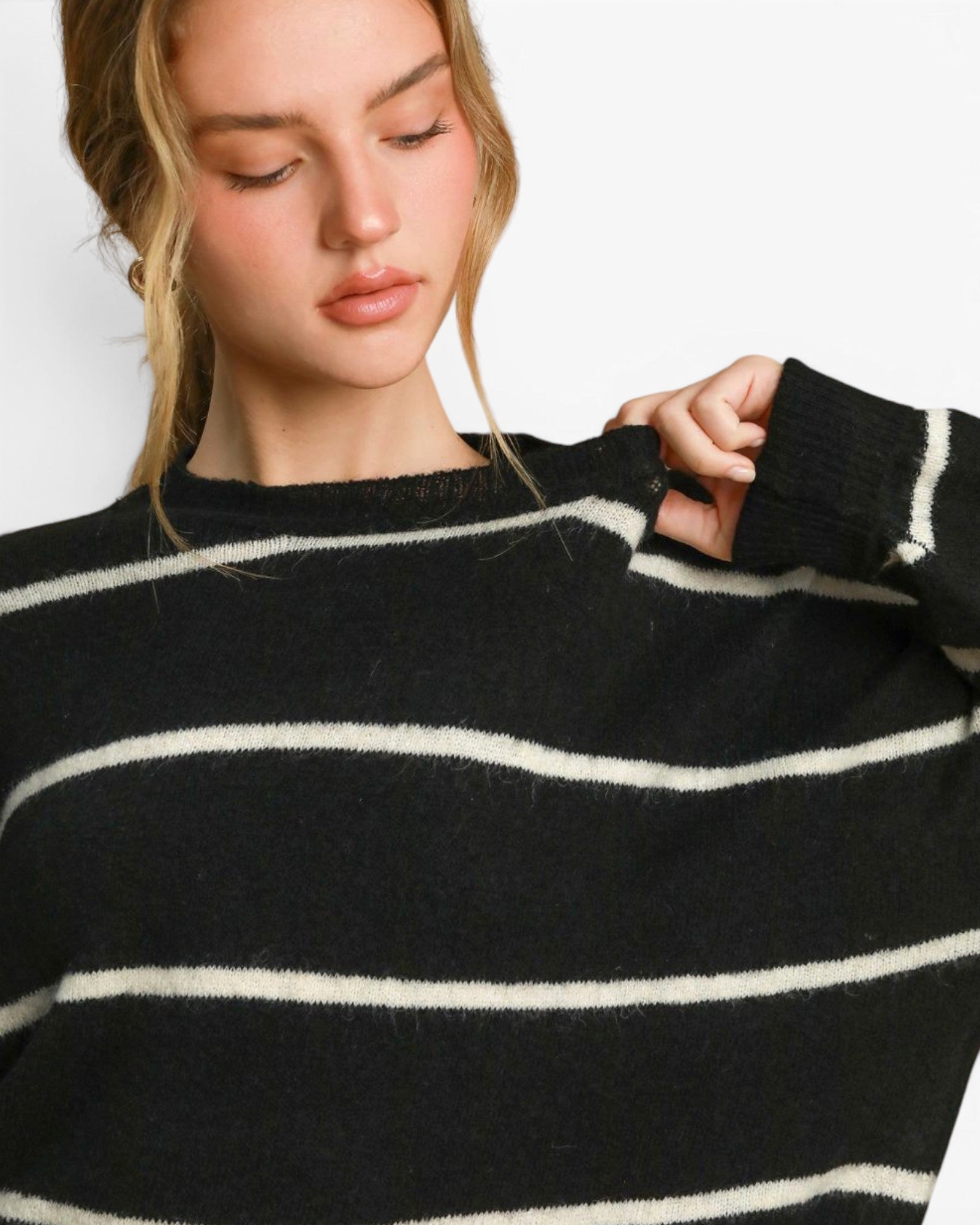 A close-up of a woman wearing a black knit sweater with horizontal white stripes, holding up the ribbed cuff of the sleeve against a plain white background.