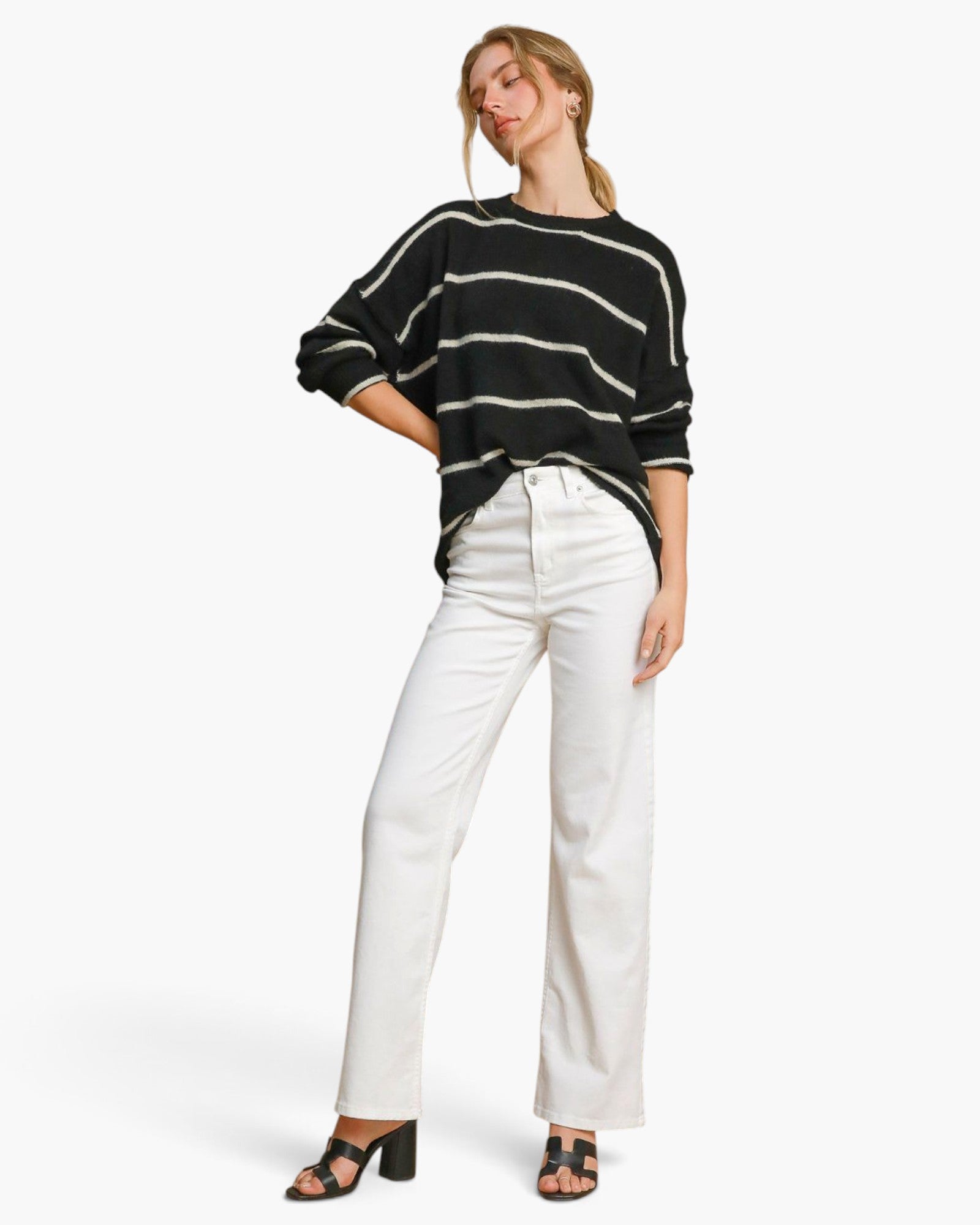 A woman modeling a black and white striped knit sweater with three-quarter sleeves, paired with white wide-leg jeans and black block-heel sandals, against a plain white studio background.
