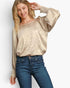A woman modeling a metallic champagne gold long-sleeve top with a textured crinkle pattern and elasticized cuffs, paired with dark blue jeans, against a light gray studio background.