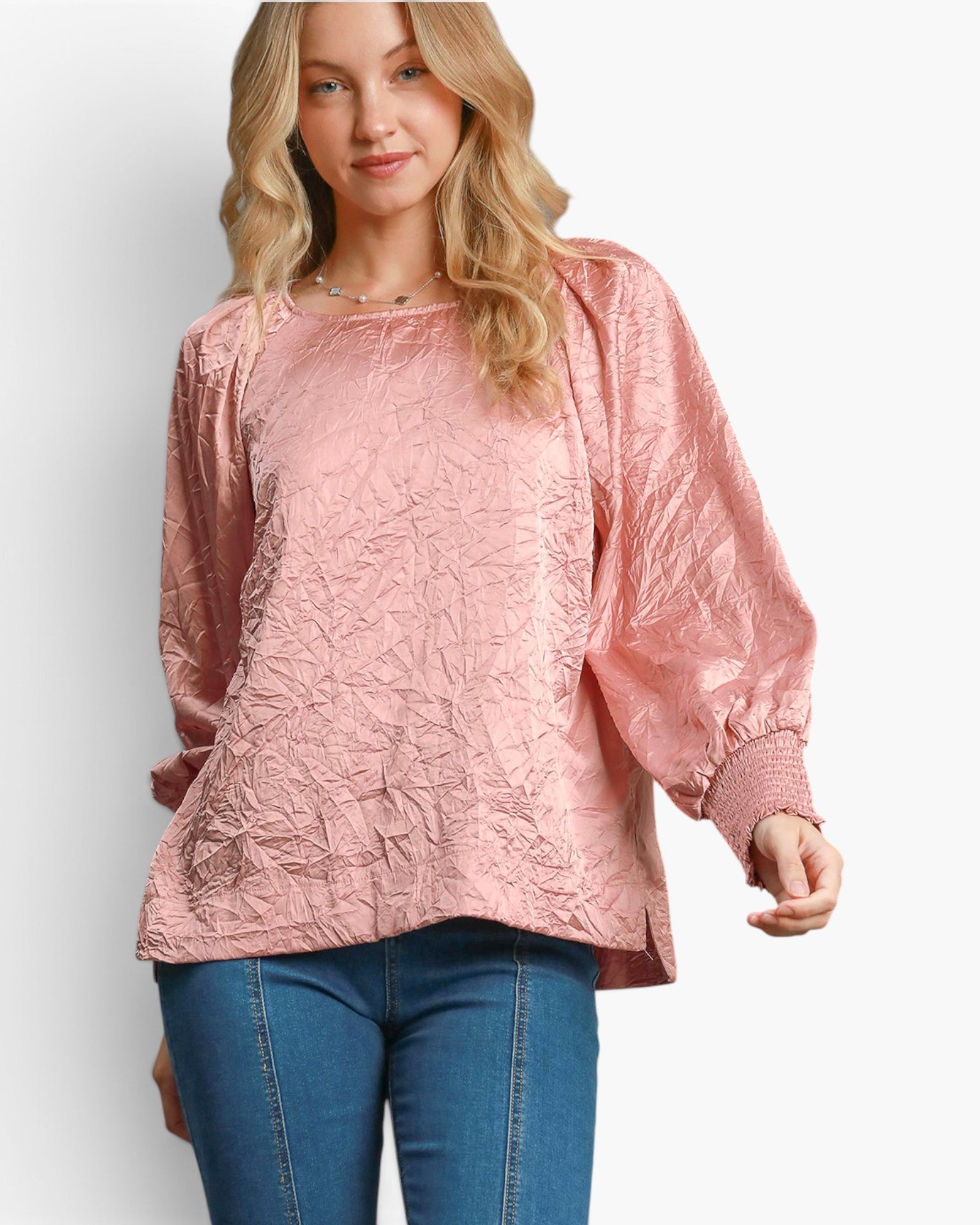 A woman wearing a dusty pink textured long-sleeve blouse with balloon sleeves and blue jeans, photographed against a light gray studio background.