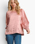 A woman wearing a dusty pink textured long-sleeve blouse with balloon sleeves and blue jeans, photographed against a light gray studio background.