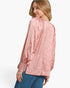 A woman wearing a textured, crinkled pink blouse with long sleeves and smocked cuffs, paired with blue jeans, shown from a three-quarters back view against a light gray studio background.