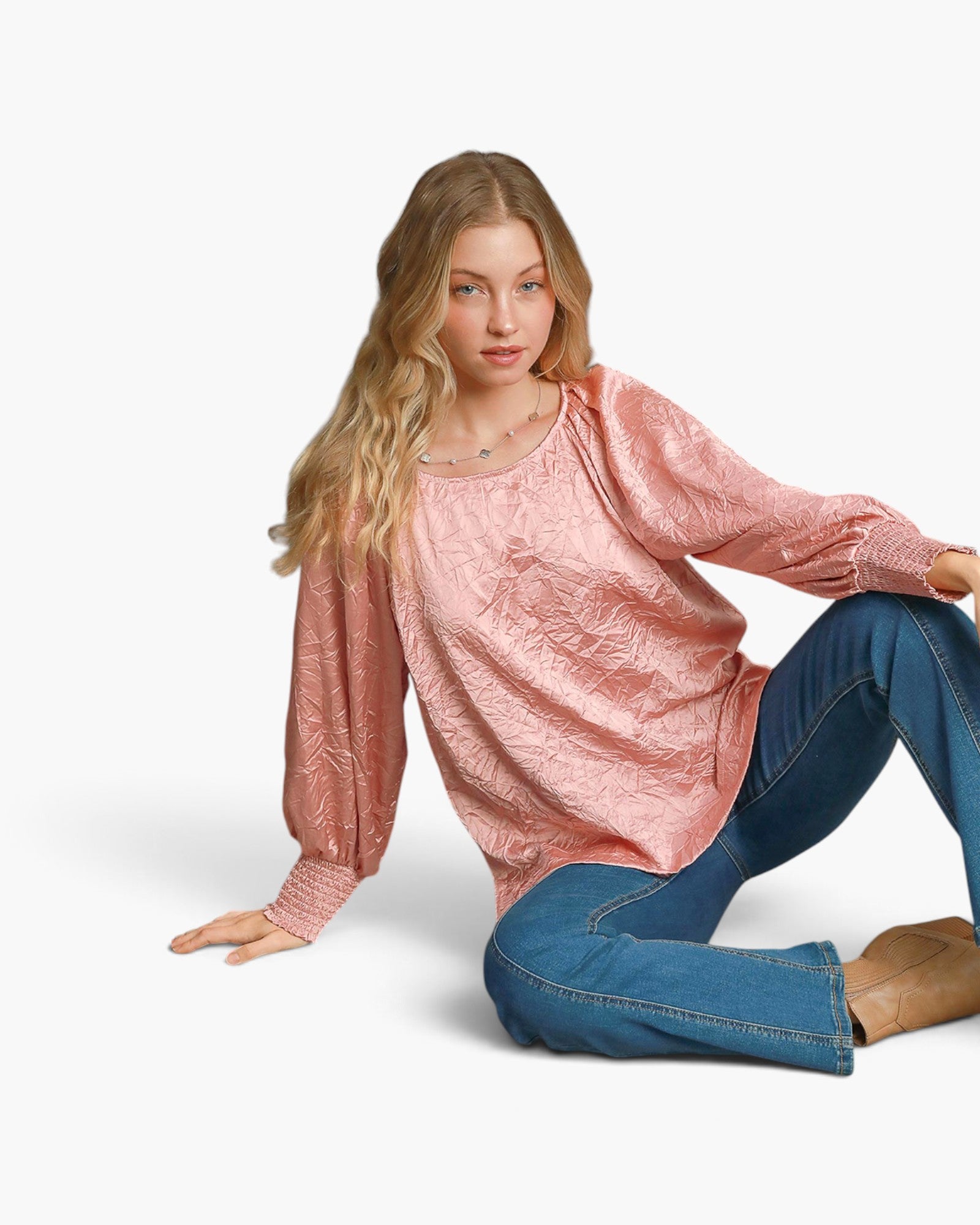 A woman sitting on a white studio background wearing a textured, metallic pink long-sleeve top and blue jeans.