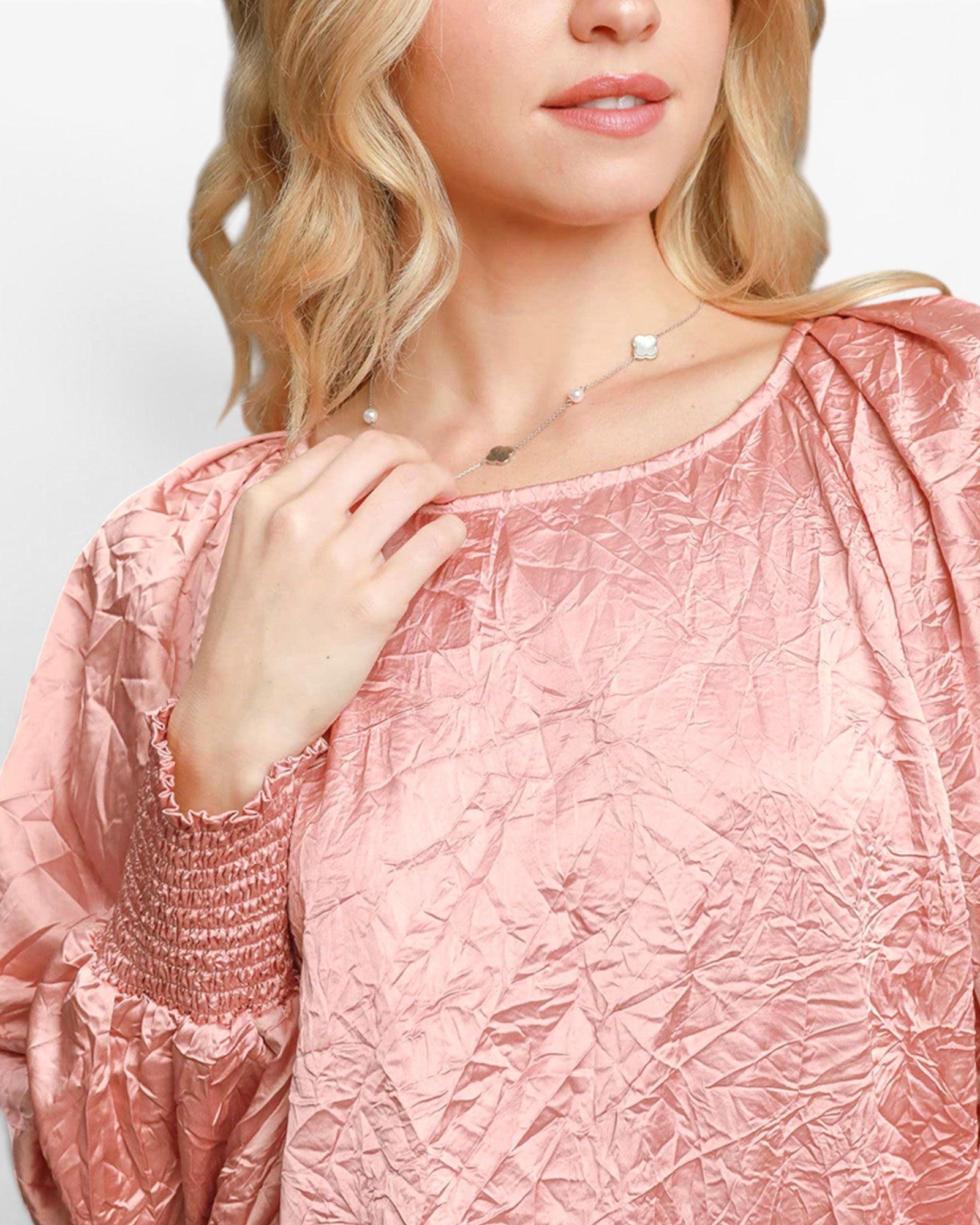 A close-up of a woman wearing a textured, shiny pink blouse with a smocked sleeve and a delicate silver necklace.