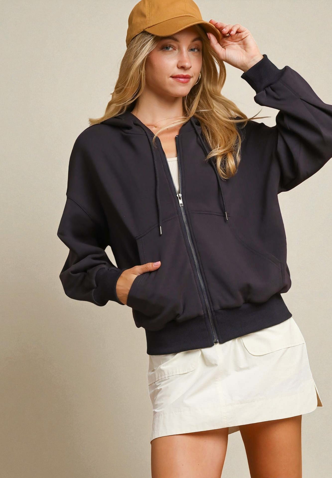 Woman wearing a black jacket, white skirt, and tan cap on a beige background