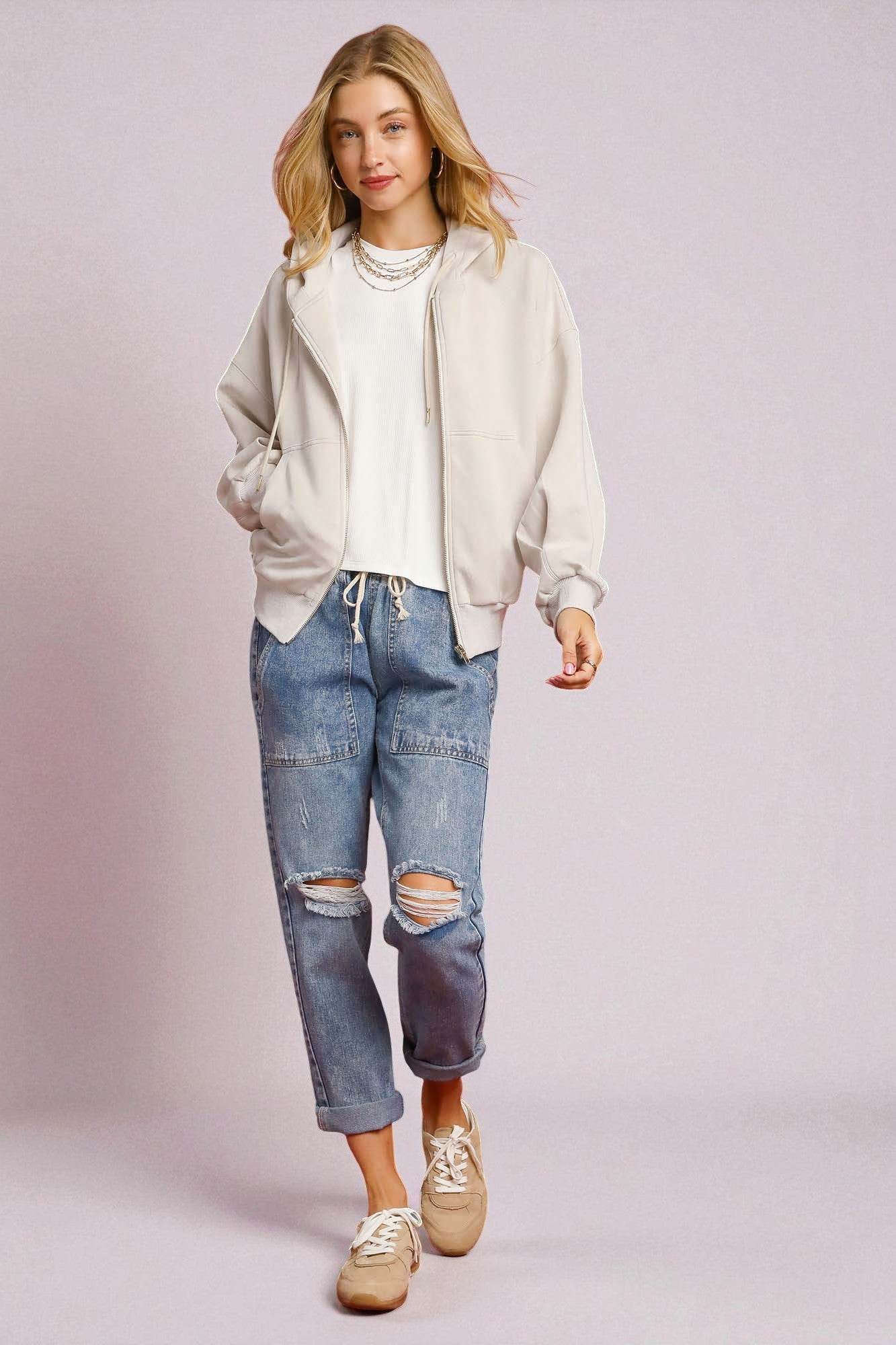 Woman wearing a cream oversized sweatshirt, white shirt, blue ripped jeans, and beige sneakers on a light pink background