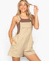 A woman modeling a beige short overall jumpsuit with a brown tank top underneath, posing against a light gray studio background.