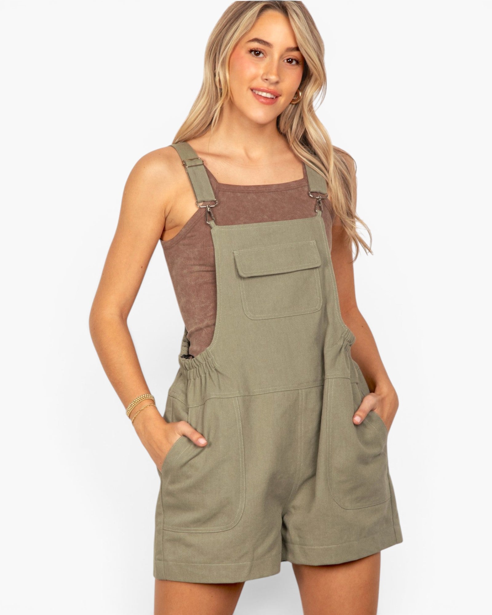 A woman modeling a pair of olive green bib shorts with a brown corduroy camisole underneath, set against a plain white studio background.