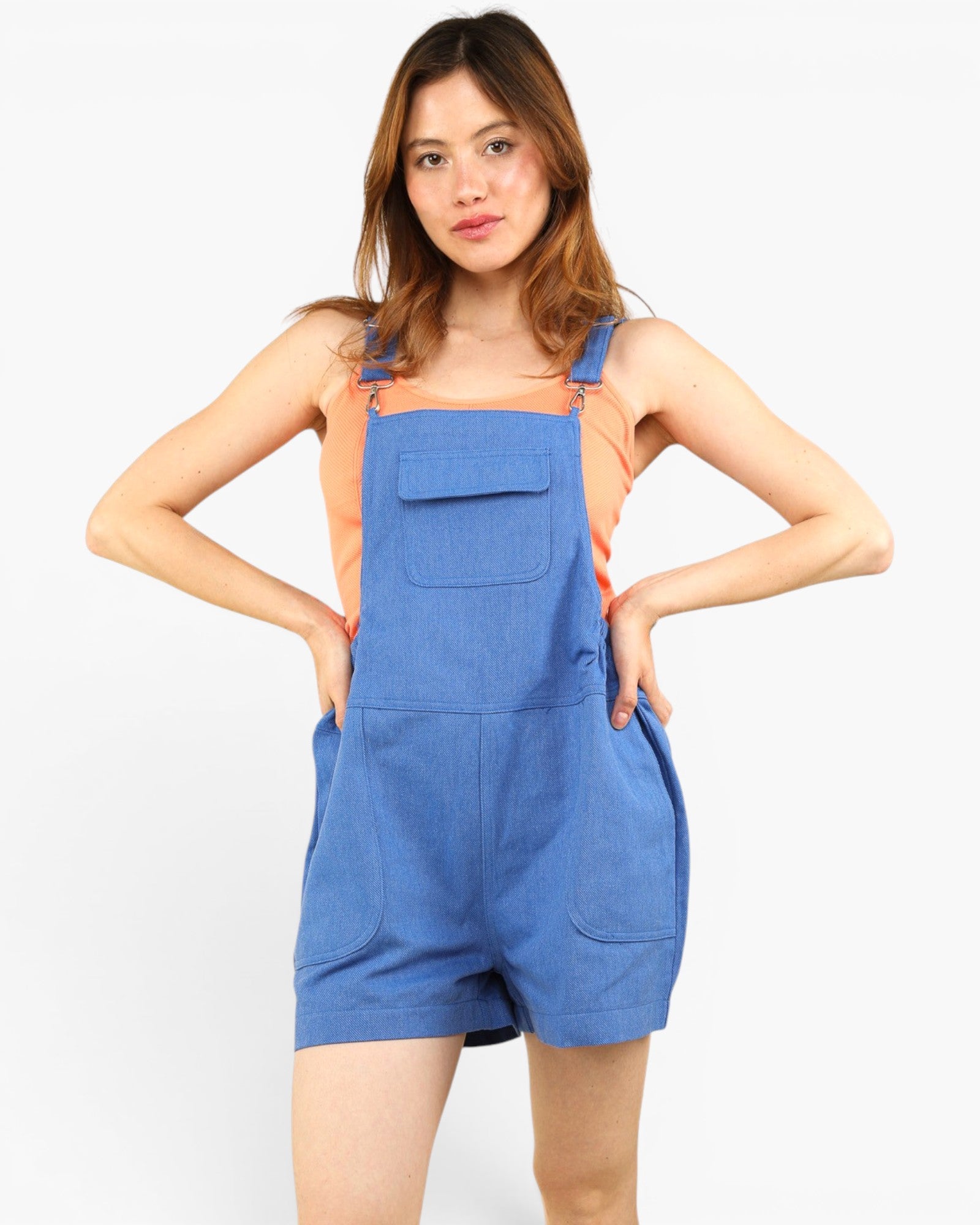A woman modeling a pair of blue denim bib shorts with shoulder straps and front pockets, worn over a peach-colored sleeveless top, against a plain white studio background.