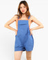 A woman modeling a pair of blue denim bib shorts with shoulder straps and front pockets, worn over a peach-colored sleeveless top, against a plain white studio background.