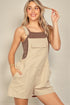 Woman wearing beige overalls with brown straps against a plain background
