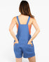 A woman shown from the back wearing a blue denim-style overall romper with shoulder straps and large back pockets, over a peach-colored sleeveless top, against a plain light gray studio background.