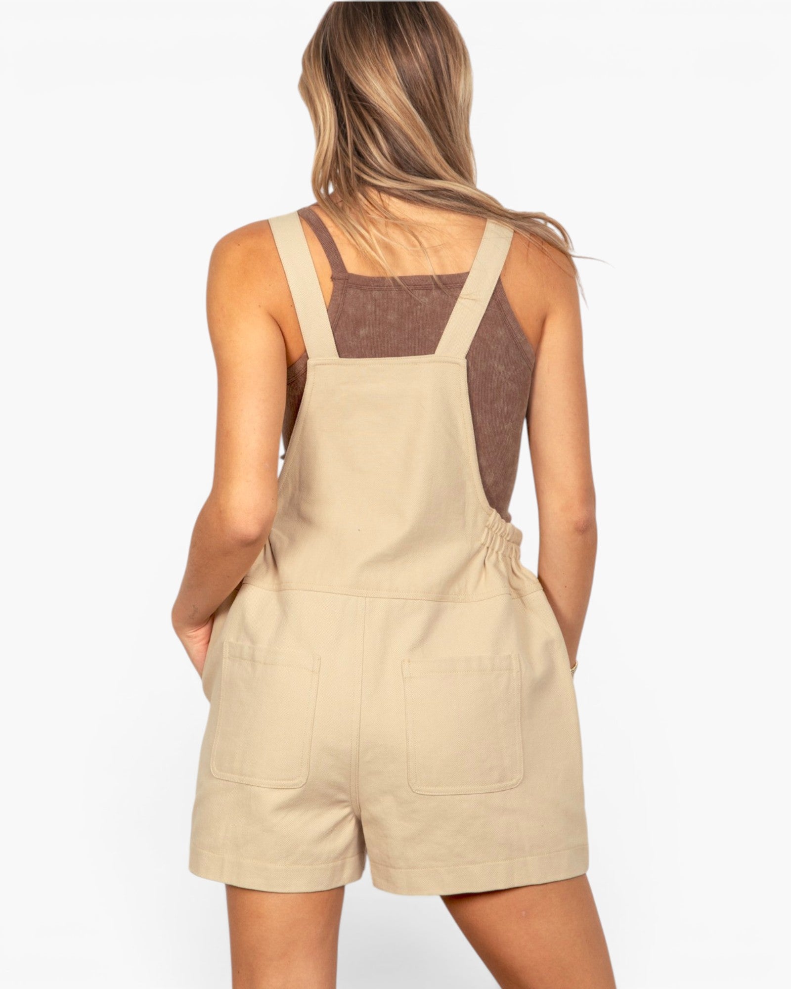 A woman shown from the back wearing a beige short overall jumpsuit over a brown tank top, against a plain white background.