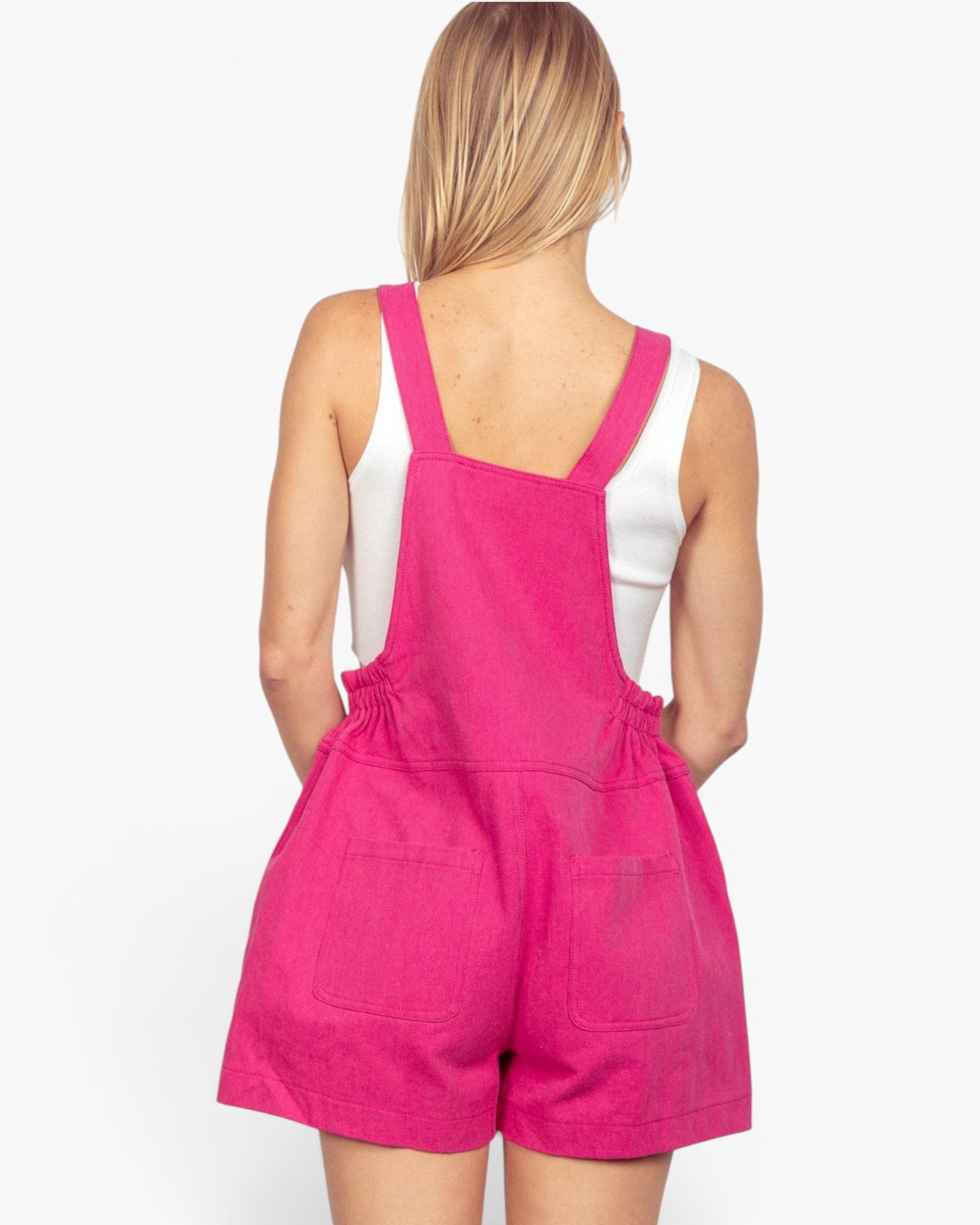 A woman shown from the back wearing bright pink denim-style overall shorts over a white tank top, against a plain light background.