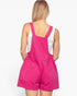 A woman shown from the back wearing bright pink denim-style overall shorts over a white tank top, against a plain light background.