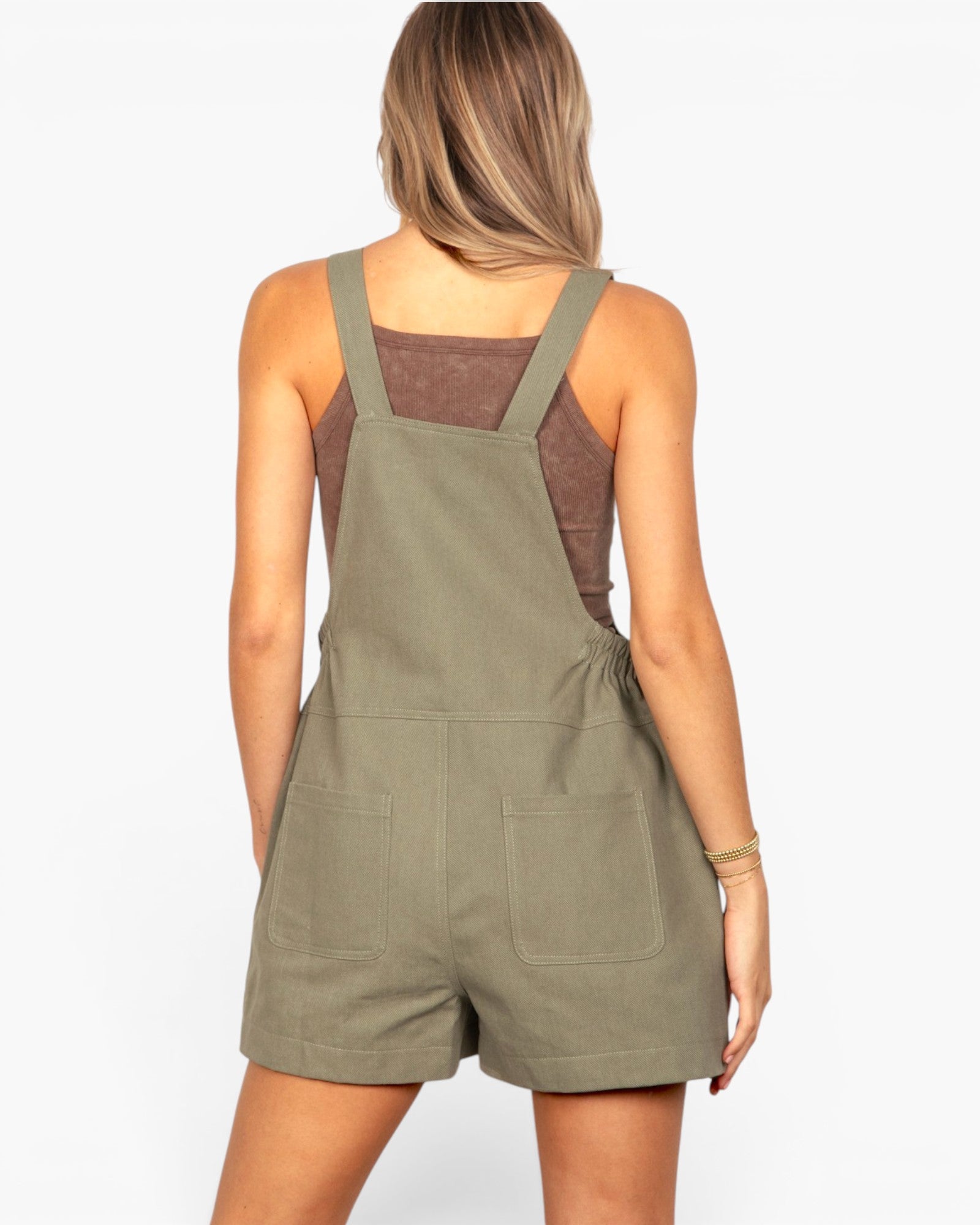 A woman shown from the back wearing an olive green short overall jumpsuit over a brown tank top, against a plain white studio background.