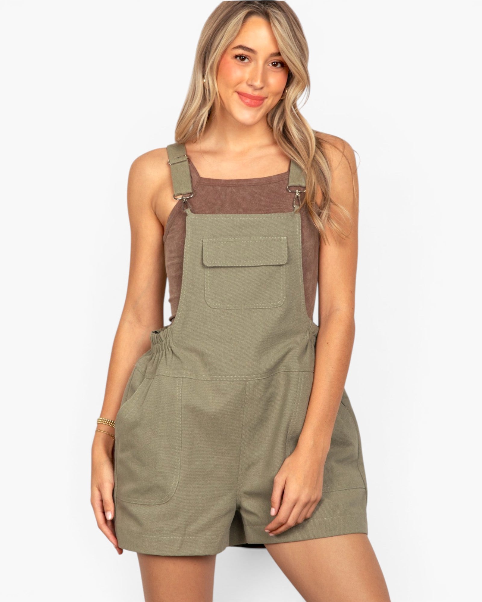 A woman modeling olive green bib shorts with a brown tank top underneath, set against a plain white studio background.