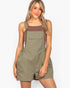 A woman modeling olive green bib shorts with a brown tank top underneath, set against a plain white studio background.