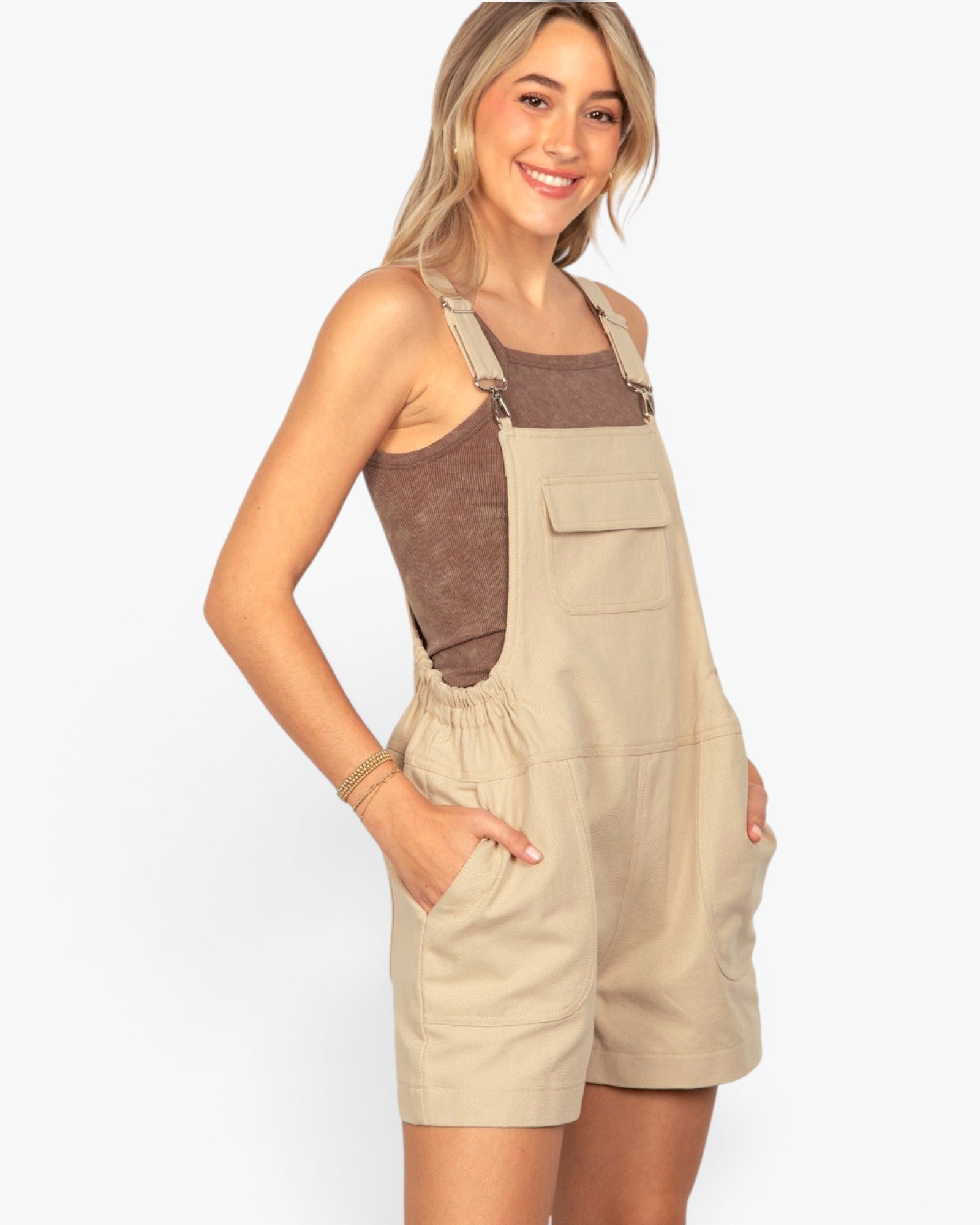 A woman modeling a pair of beige utility bib shorts with a brown textured tank top underneath, set against a plain white studio background.