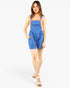 A woman modeling a pair of blue overall shorts with shoulder straps and side pockets, paired with white slide sandals, against a plain light gray studio background.