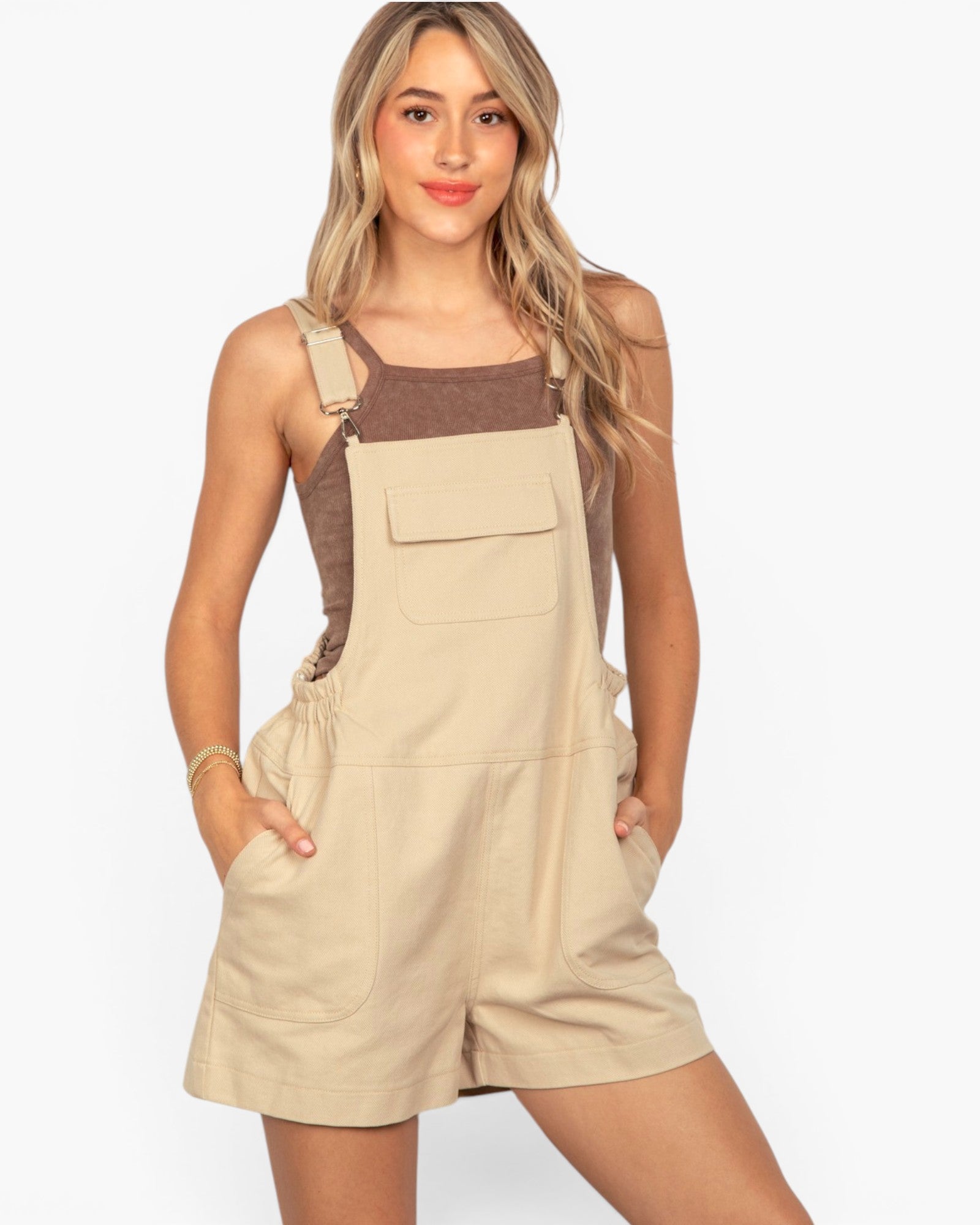 A woman modeling a beige short bib overalls romper with a brown knit tank top underneath, set against a plain light gray studio background.