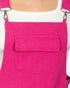 Bright pink denim-style bib overalls with a front flap pocket, shown worn over a white top. The straps are visible at the top corners with metal clips.
